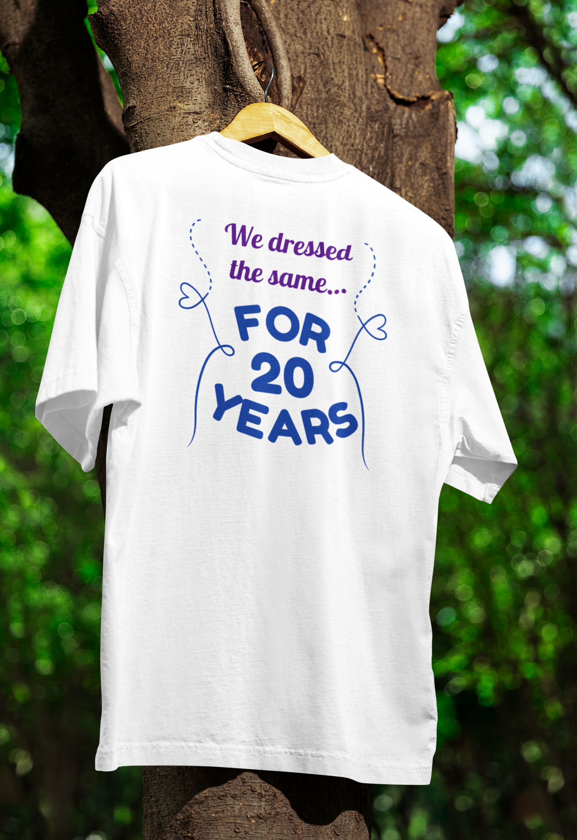 White t-shirt for Twins with text 'We dressed the same... FOR 20 YEARS' on a wooden hanger against a tree and green foliage background.